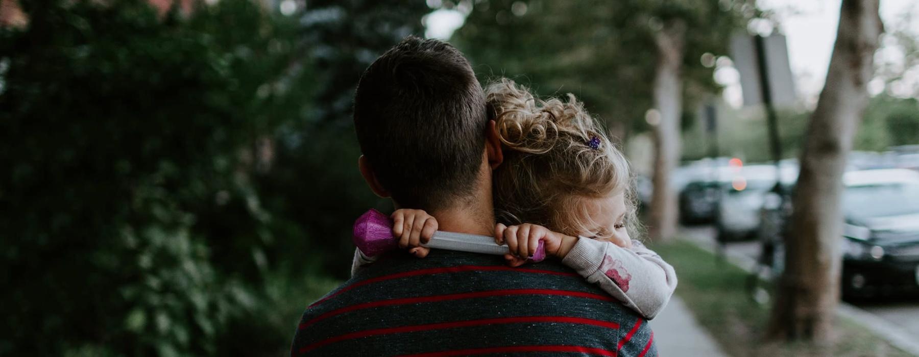 tired little girl rests her head on her father's shoulder as he carries her down the city sidewalk