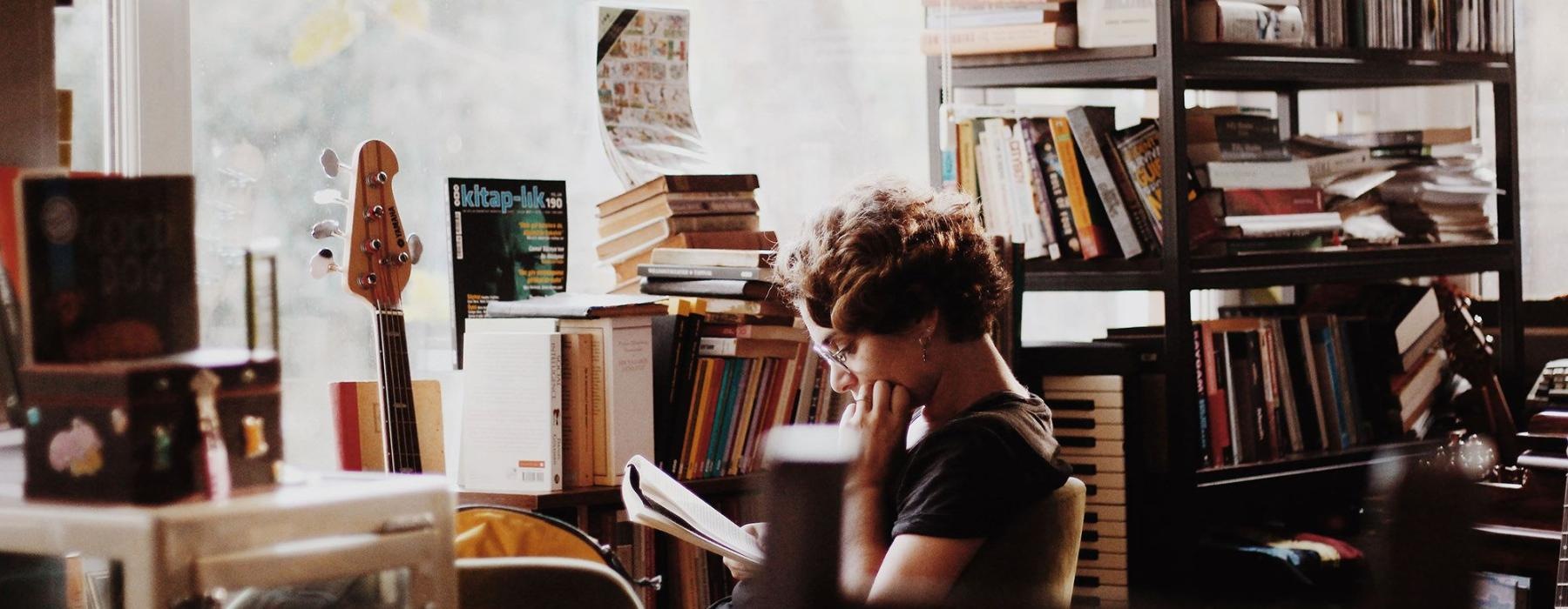 woman, surrounded by books, sits and reads in front of a picture window